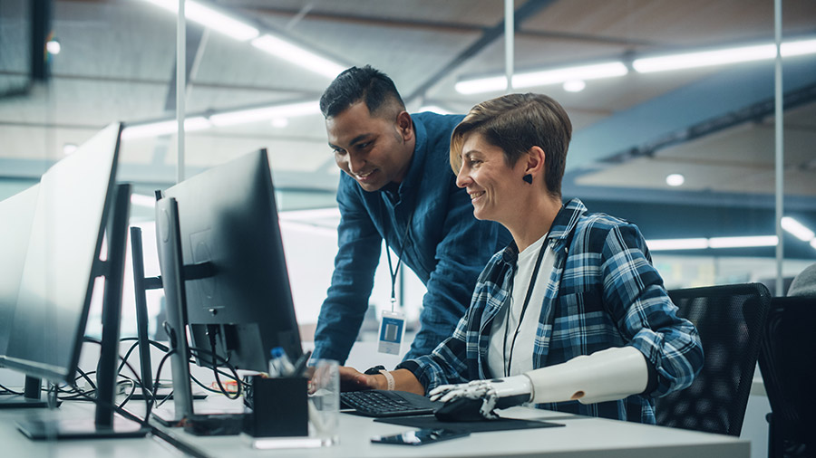 Image of woman with a prosthetic arm and hand working at a computer talking to a male colleague