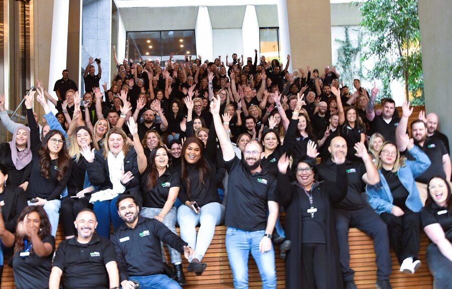 A large group of people in black polo shirts with Jobfind logo wave and smile at the camera.