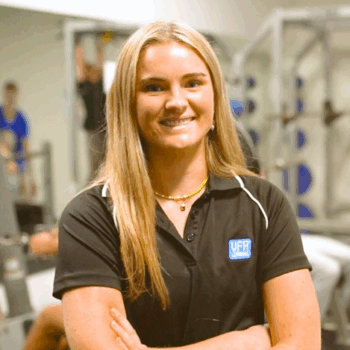Hannah with long blonde hair, wearing a brown gym shirt smiles into camera with crossed arms.