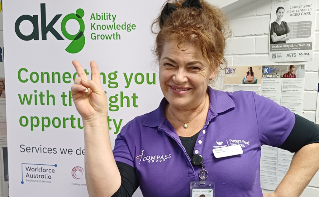 A smiling woman named Amal in a purple Western Health Patient Food Services uniform, giving a peace sign in front of an akg (Ability Knowledge Growth) banner