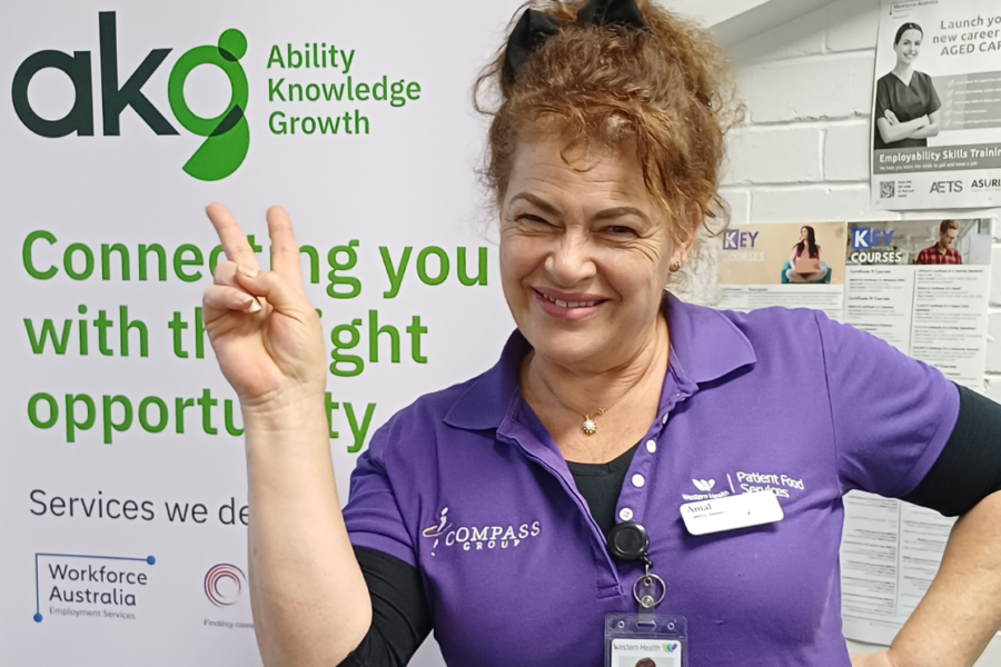 A smiling woman named Amal in a purple Western Health Patient Food Services uniform, giving a peace sign in front of an akg (Ability Knowledge Growth) banner