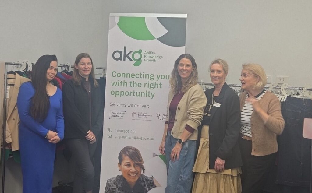 Five women posing together next to an AKG (Ability Knowledge Growth) banner indoors, surrounded by clothing racks filled with professional attire.