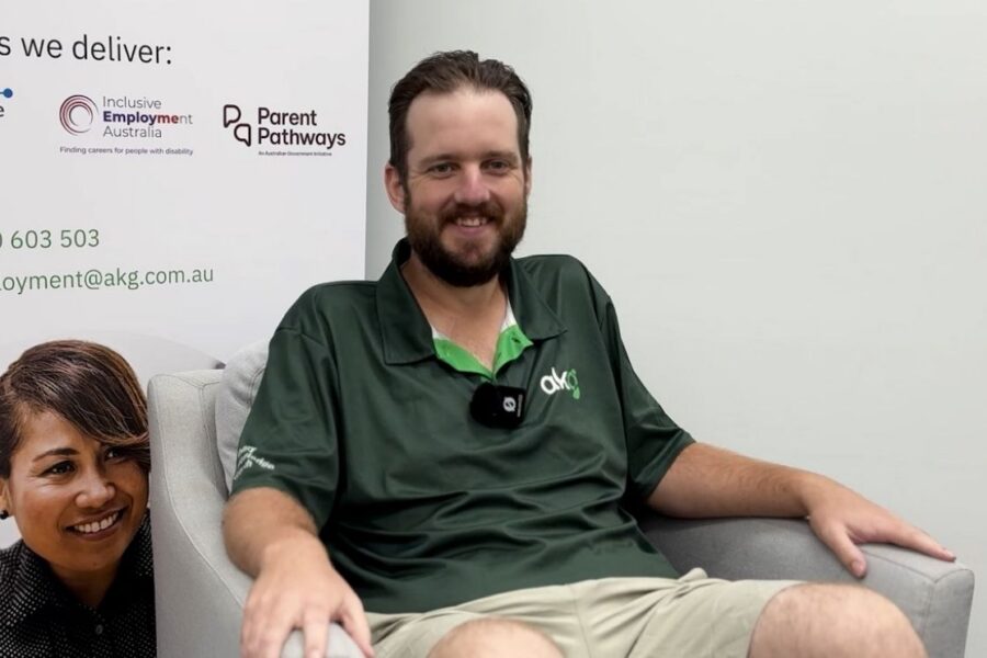 Tristan, an AKG Job Coach, smiling while seated in an armchair indoors, wearing a green AKG polo shirt with a lapel microphone attached.