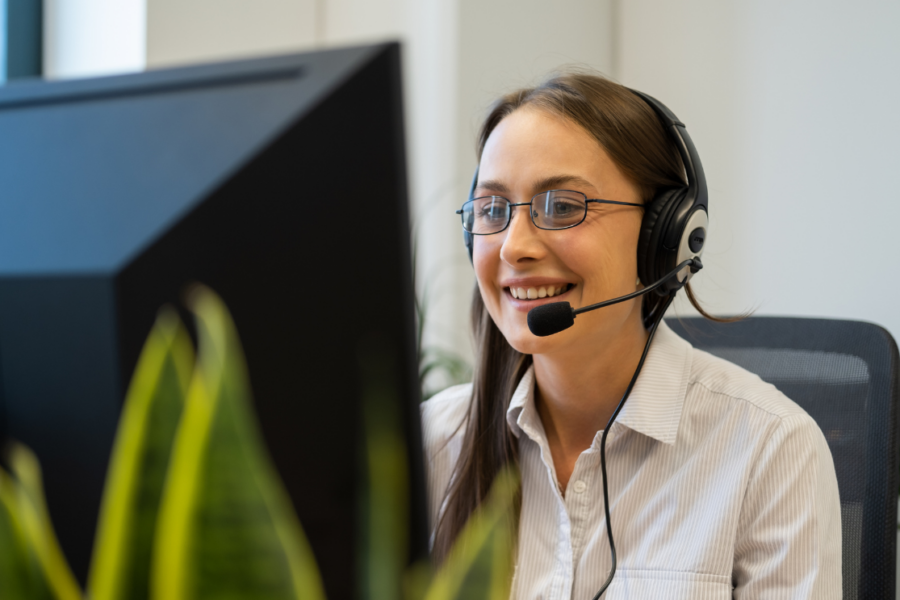 Smiling customer support representative wearing a headset with a microphone, sitting at a desk and looking at a computer monitor