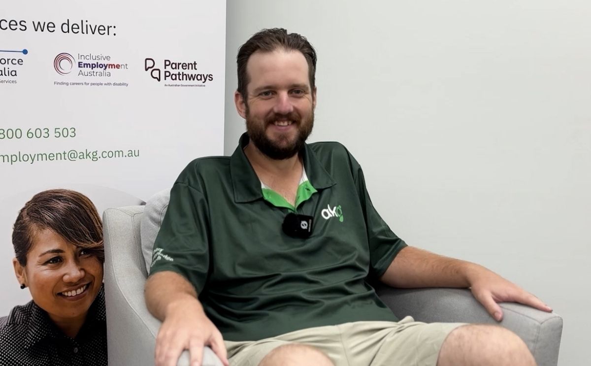 Tristan, an AKG Job Coach, smiling while seated in an armchair indoors, wearing a green AKG polo shirt with a lapel microphone attached.
