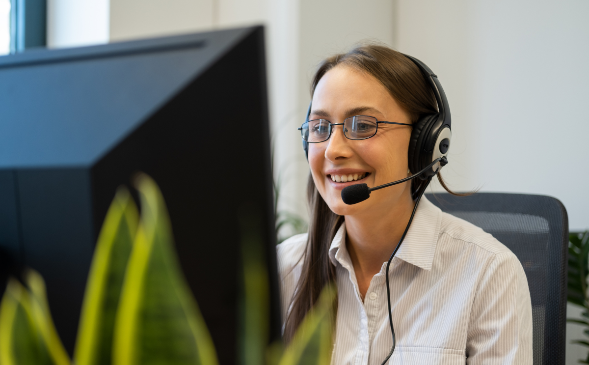 Smiling customer support representative wearing a headset with a microphone, sitting at a desk and looking at a computer monitor