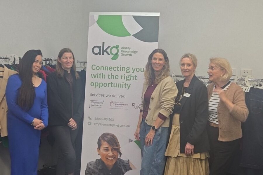 Five women posing together next to an AKG (Ability Knowledge Growth) banner indoors, surrounded by clothing racks filled with professional attire.