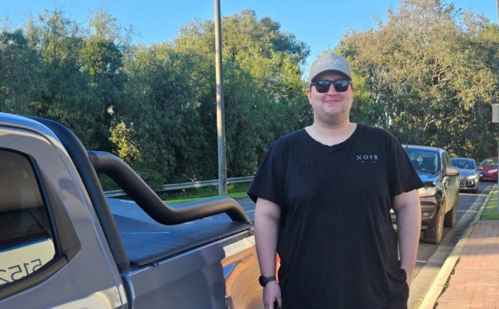Tyler standing outdoors beside a pickup truck on a sunny day, wearing a black t-shirt, sunglasses, and a grey cap.