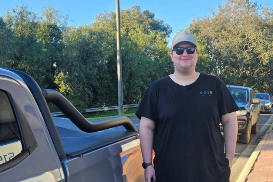 Tyler standing outdoors beside a pickup truck on a sunny day, wearing a black t-shirt, sunglasses, and a grey cap.