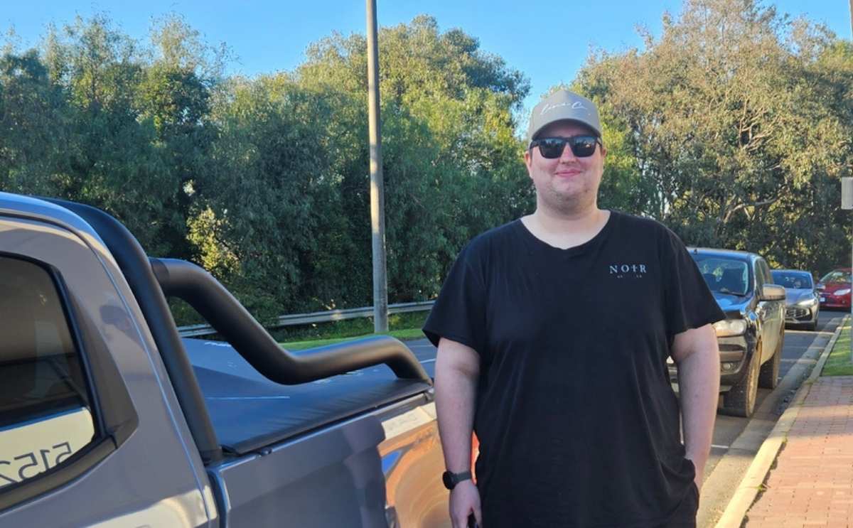 Tyler standing outdoors beside a pickup truck on a sunny day, wearing a black t-shirt, sunglasses, and a grey cap.