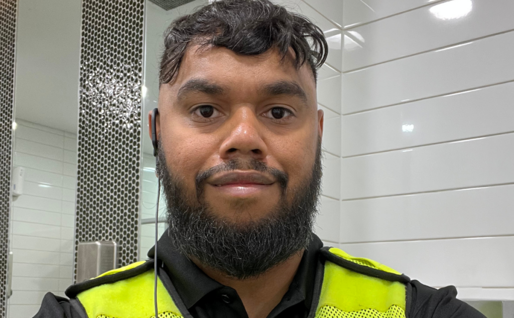 Ngudu wearing a high-visibility work vest indoors against a white-tiled wall
