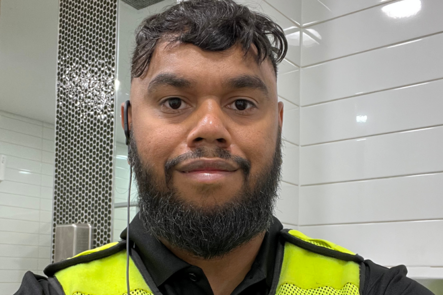 Ngudu wearing a high-visibility work vest indoors against a white-tiled wall