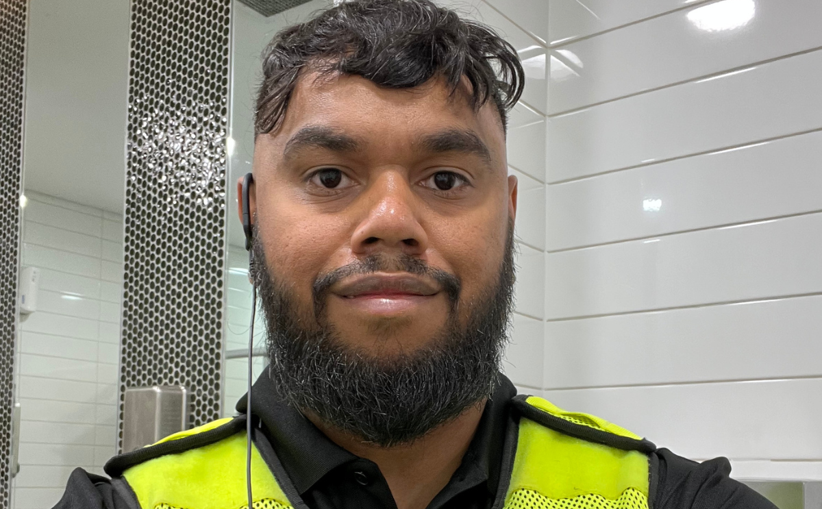Ngudu wearing a high-visibility work vest indoors against a white-tiled wall