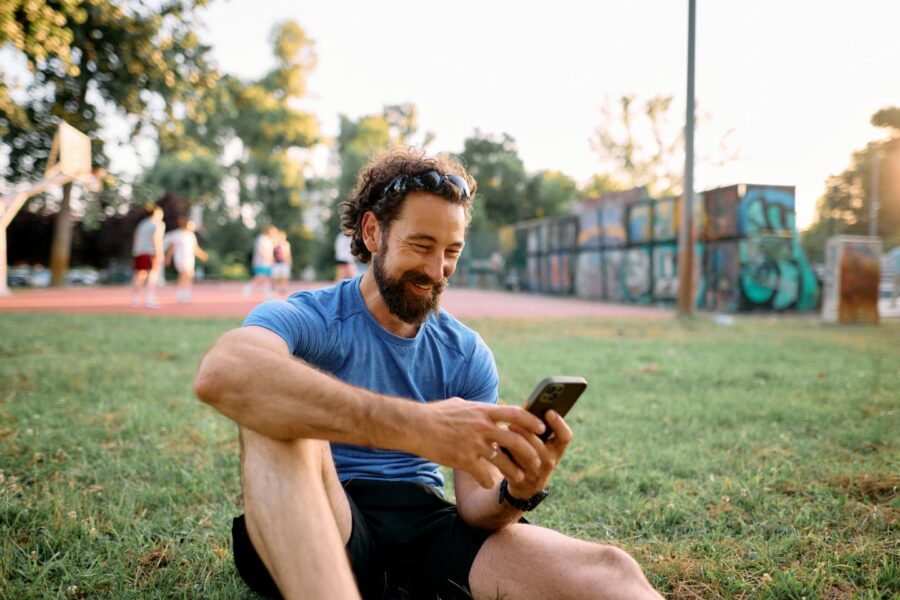Happy sportsman sitting on the grass in a park checking his smartphone after training