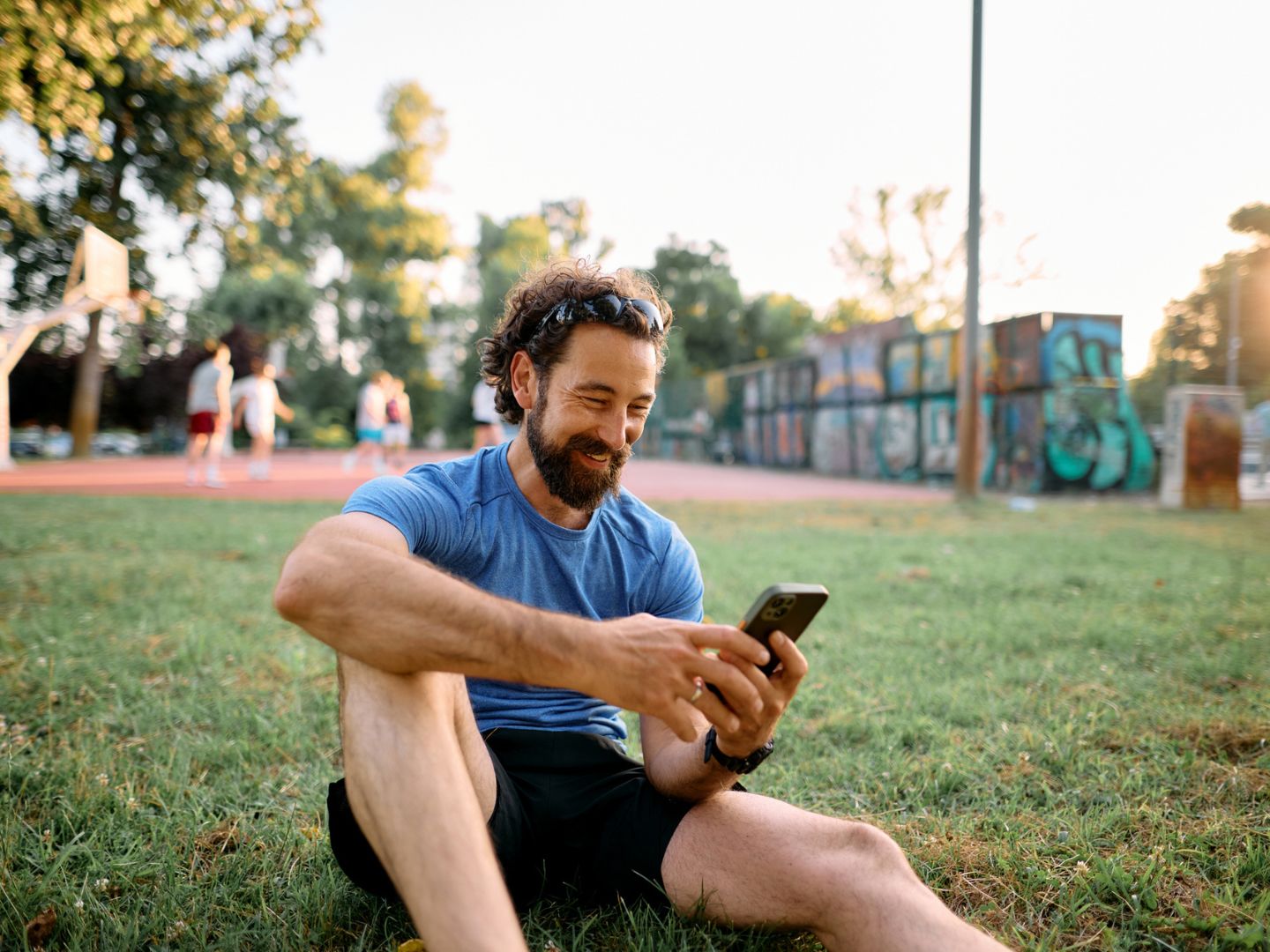Happy sportsman sitting on the grass in a park checking his smartphone after training