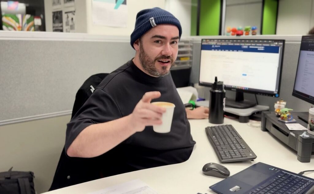 Man in a beanie holding a small takeaway coffee cup during a barista training session at a computer desk.