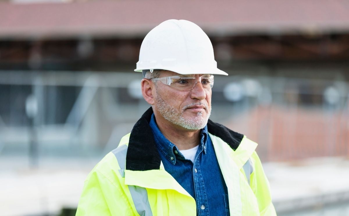 Male construction worker or site manager wearing a white hard hat, safety glasses, and high-visibility jacket.