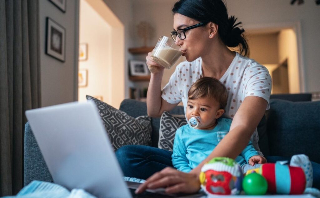 A mother holding her toddler on her lap while drinking coffee and working on a laptop.