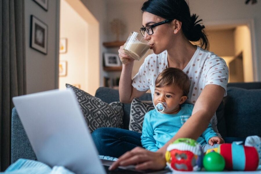 A mother holding her toddler on her lap while drinking coffee and working on a laptop.