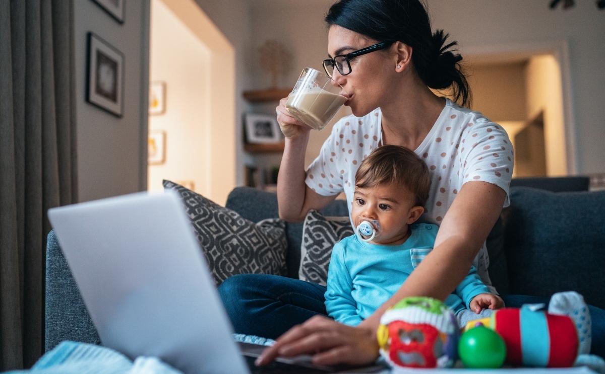 A mother holding her toddler on her lap while drinking coffee and working on a laptop.