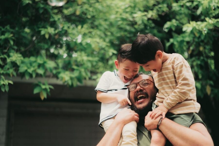 A smiling father carrying his two young sons on his shoulders outdoors under lush green trees.