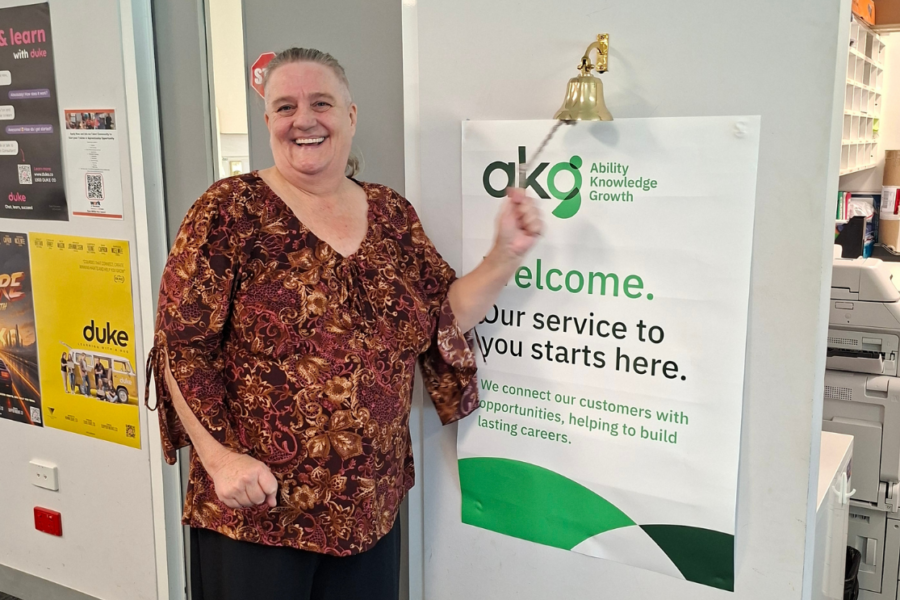 Jacqui smiling as she rings a brass success bell mounted next to an AKG poster that reads "Welcome. Our service to you starts here.