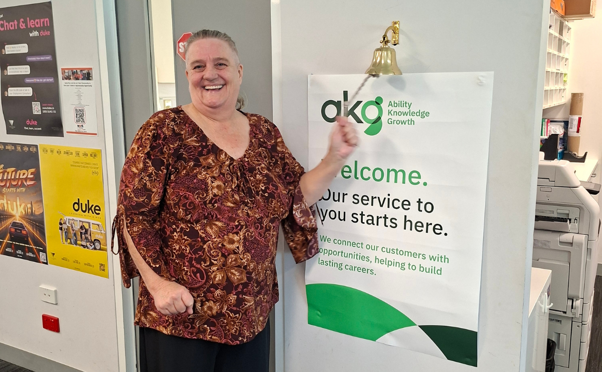 Jacqui smiling as she rings a brass success bell mounted next to an AKG poster that reads "Welcome. Our service to you starts here.