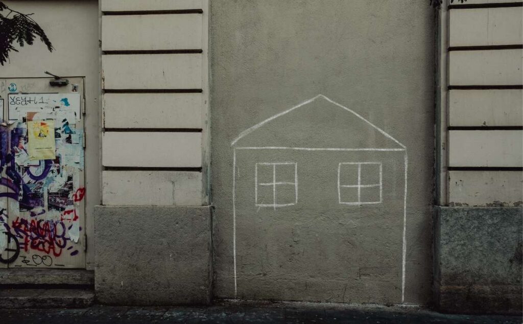A outline of a house drawn in white chalk on an urban concrete wall next to a door covered in graffiti.