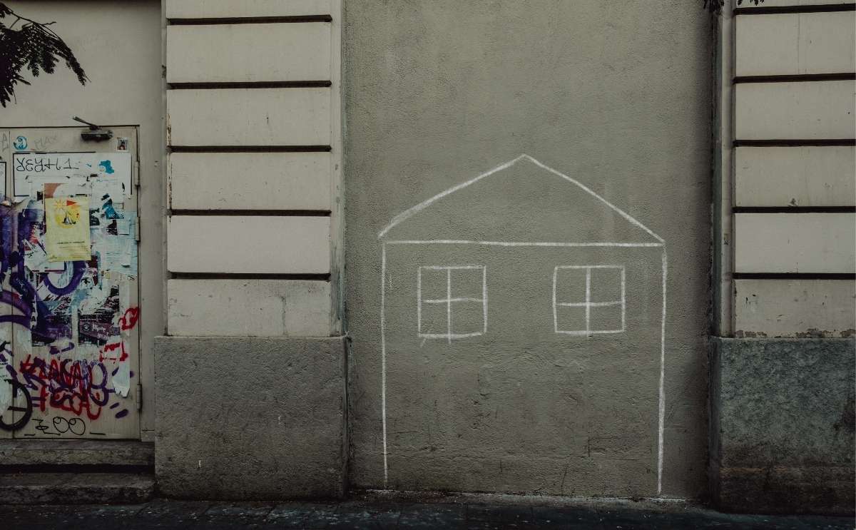 A outline of a house drawn in white chalk on an urban concrete wall next to a door covered in graffiti.