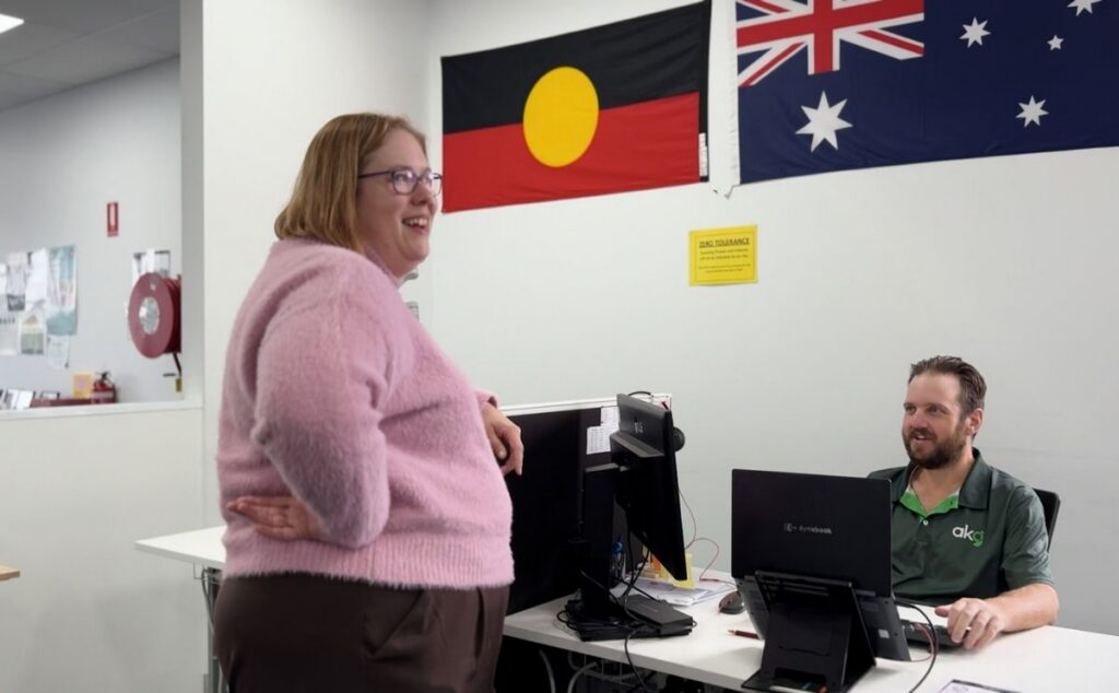A smiling woman talking with an AKG team member at a office desk with Australian and Aboriginal flags in the background.